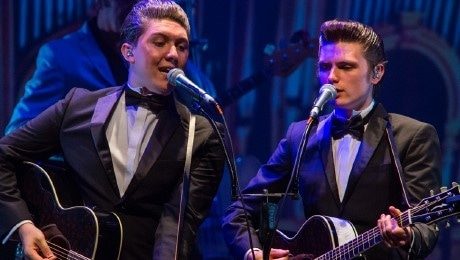 Walk Right Back Two men in tuxedos singing into microphones while playing acoustic guitars on stage.