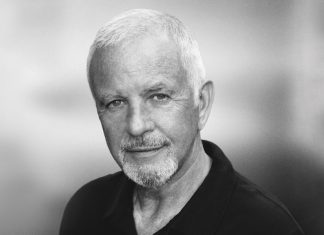 Older man with short gray hair and a mustache, wearing a black shirt against a blurred background.