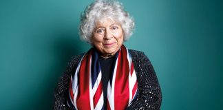 Miriam Margolyes London 2026-photo of Miriam Margolyes, draped in union jack flag on a green background smiling at the camera