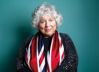 Miriam Margolyes London 2026-photo of Miriam Margolyes, draped in union jack flag on a green background smiling at the camera